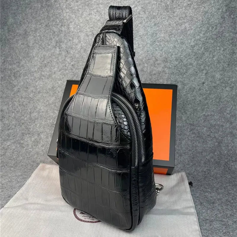 A glossy black crocodile-embossed sling bag with a top zipper, standing upright on a white dust bag with its black and orange brand box behind it against a gray textured backdrop.