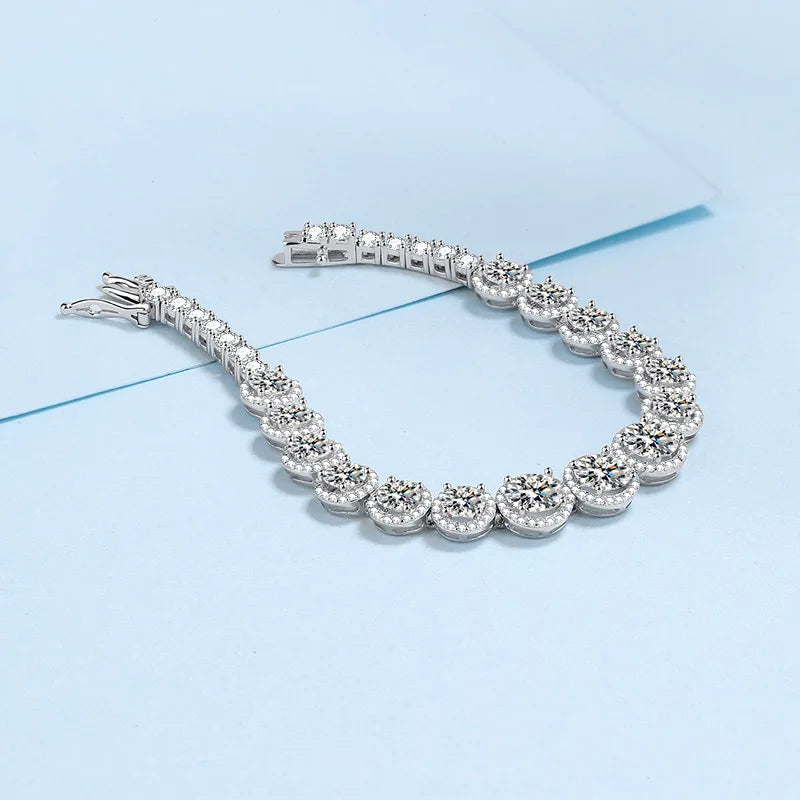 A silver tennis bracelet with round-cut clear stones, arranged in a curved shape on a light blue background.