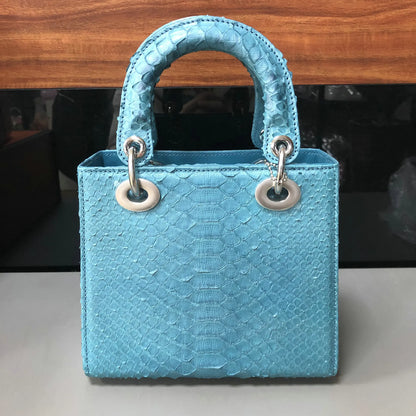 A light blue python-textured leather handbag with a top handle and silver-tone hardware, photographed on a light surface with a dark background.