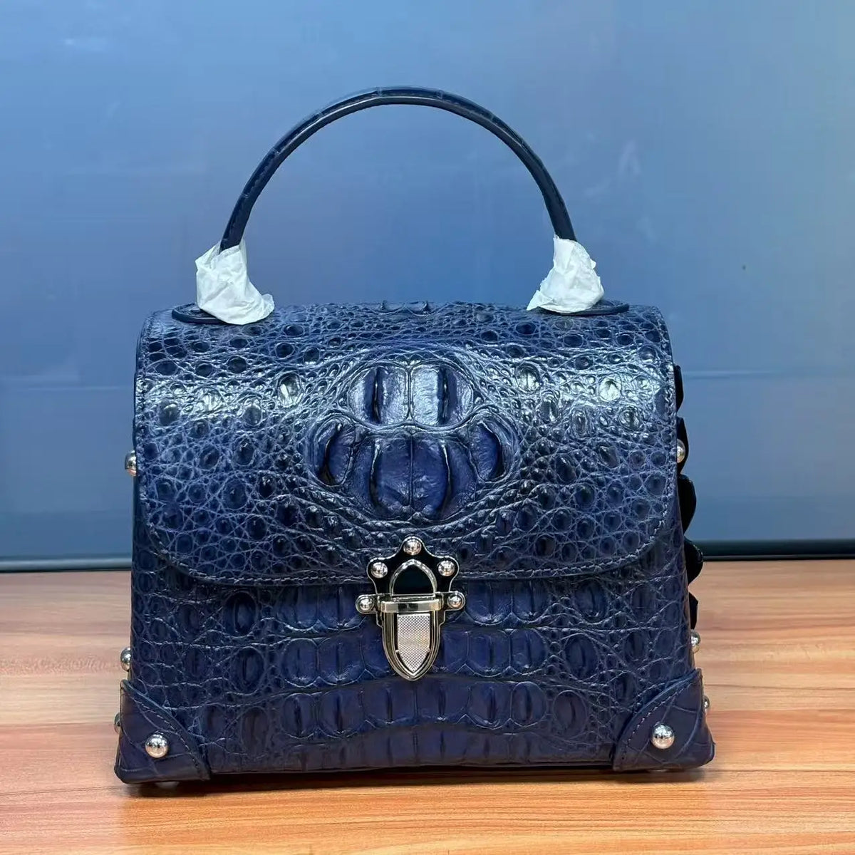 A dark blue crocodile-embossed leather handbag with a top handle, silver-tone studs, and a decorative metal clasp, shown on a wooden surface against a blue background.