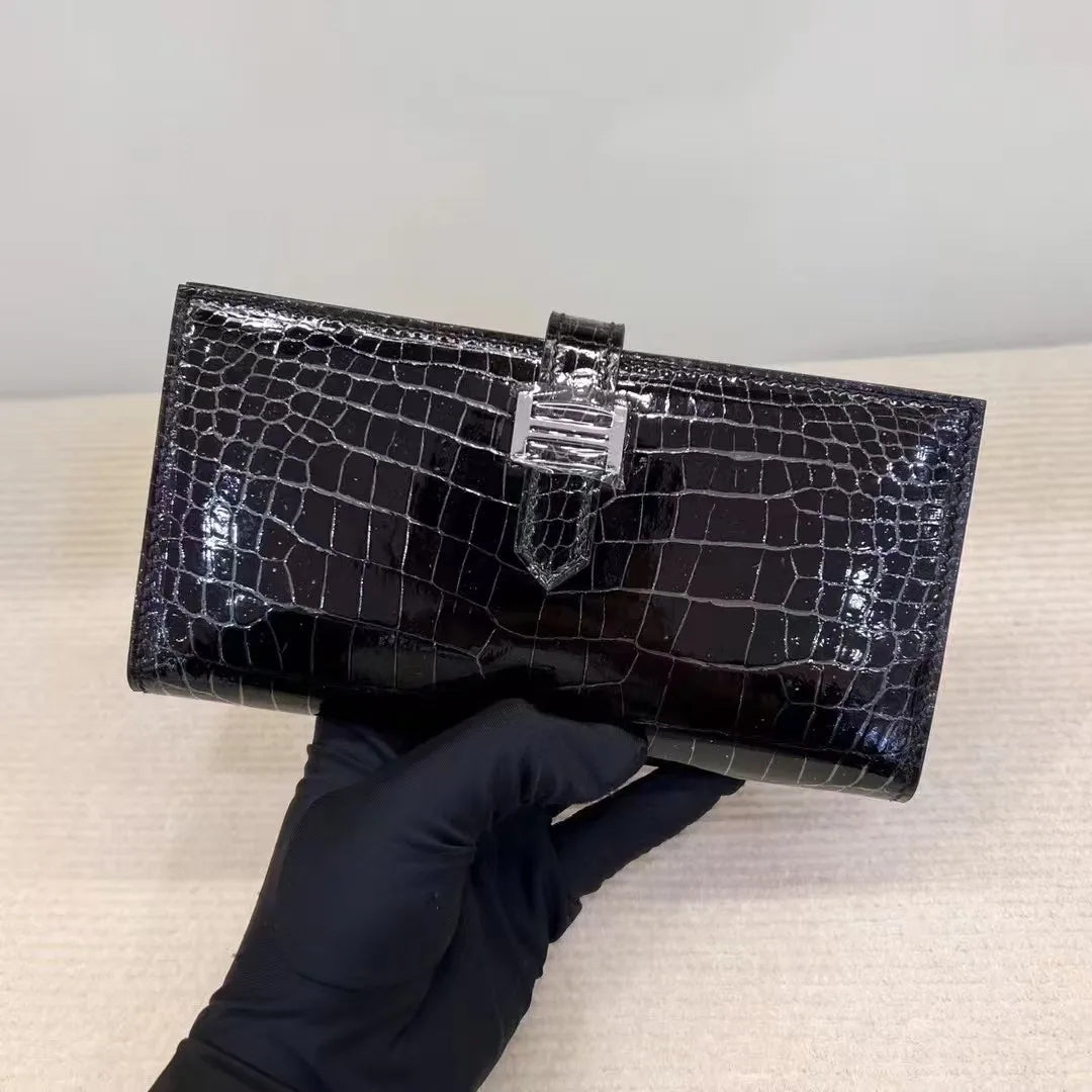 A glossy black crocodile-embossed clutch wallet held by a black gloved hand against a neutral beige background.