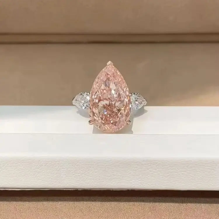 A pear-cut pink diamond ring with a silver-toned band, displayed on a white jewelry box against a beige background.
