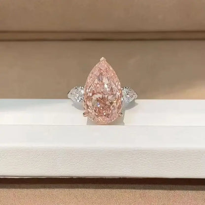 A pear-cut pink diamond ring with a silver-toned band, displayed on a white jewelry box against a beige background.