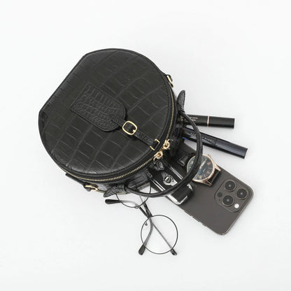 A black crocodile-embossed zippered pouch bag with gold hardware, styled with various everyday accessories including a smartphone, eyeglasses, a watch, and pens on a clean white background.