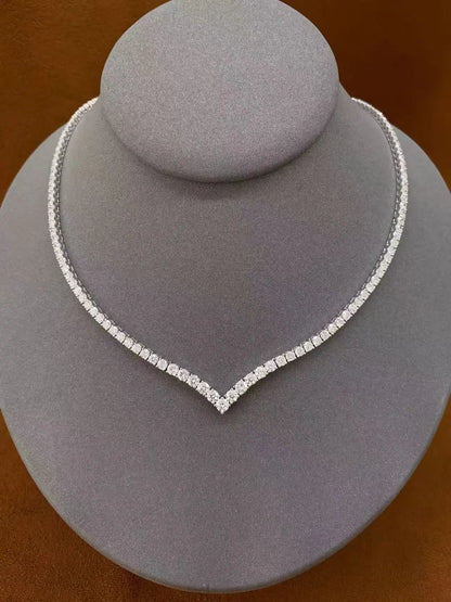 A silver-toned tennis-style necklace with a V-shaped center, displayed on a gray jewelry bust against a warm brown background.