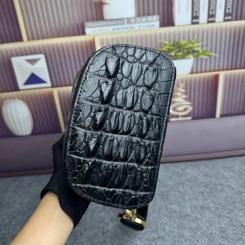 A black crocodile-embossed leather handbag held by a hand wearing a black glove, set against a blurred background of a wooden shelf and books.