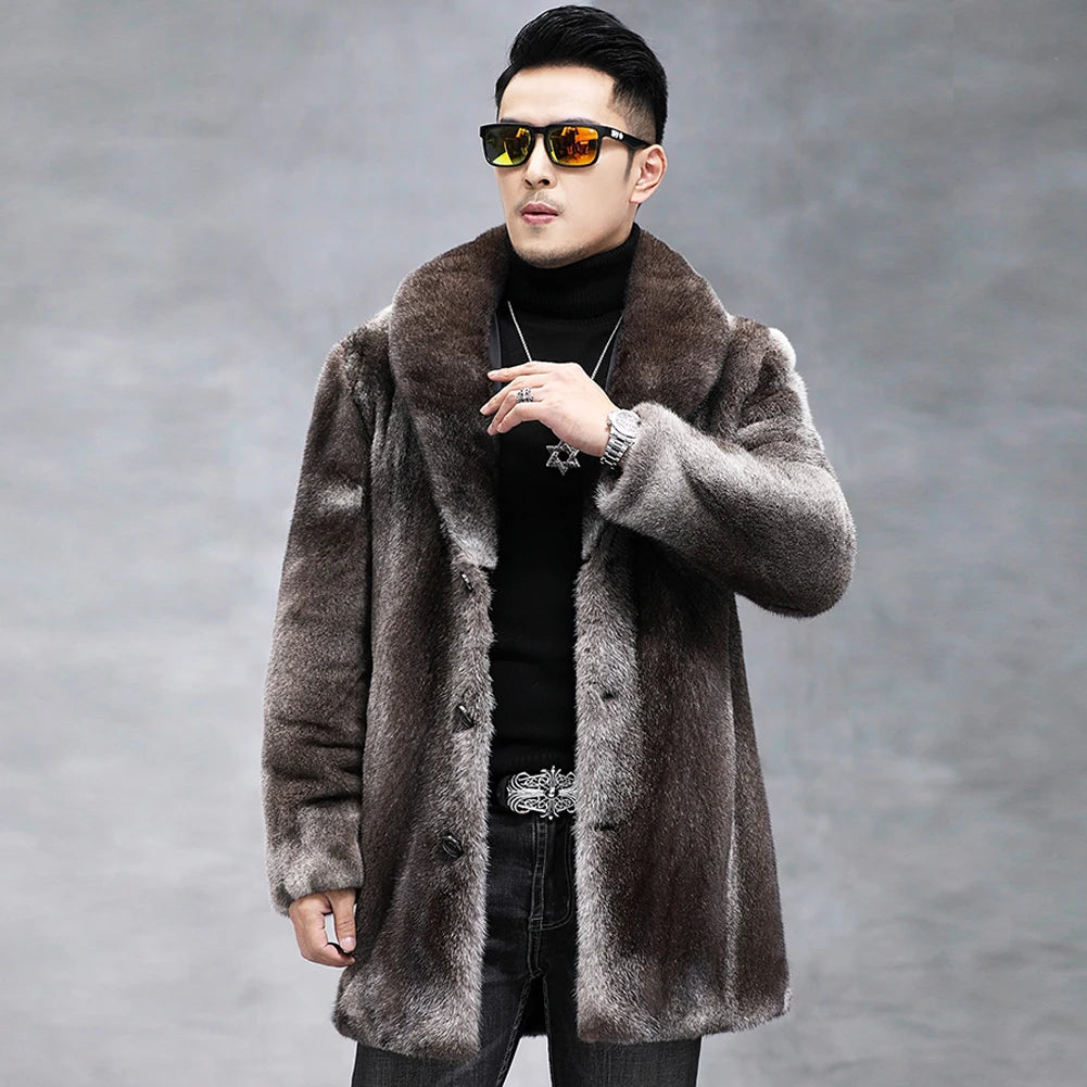 A man wearing a plush brown faux fur coat over a black turtleneck sweater and dark jeans, styled with sunglasses, a silver necklace, and a silver belt, set against a gray studio background.