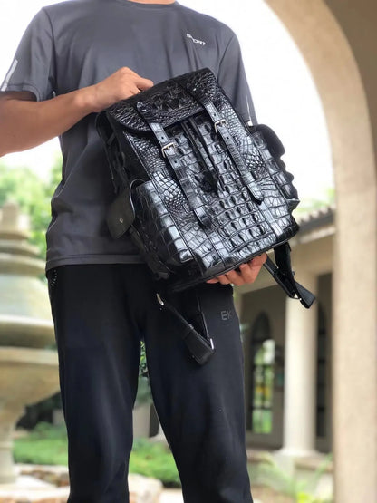 A person wearing a dark gray t-shirt and black sweatpants holds a black crocodile-embossed leather backpack in front of an outdoor stone archway.