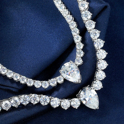 A close-up of a silver-toned necklace with clear teardrop-shaped center stones and rows of round clear stones, draped on dark navy blue fabric.
