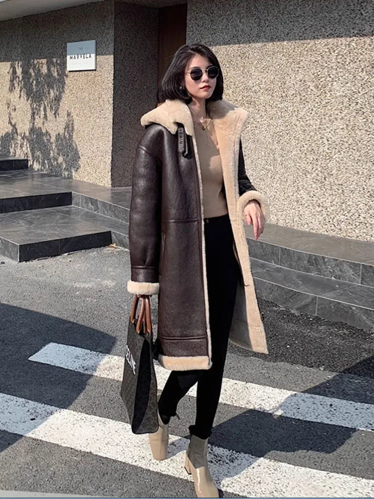 A woman wearing a dark brown shearling-lined faux-leather coat with beige trim, a beige top, black pants, and beige ankle boots, standing outdoors on a crosswalk.