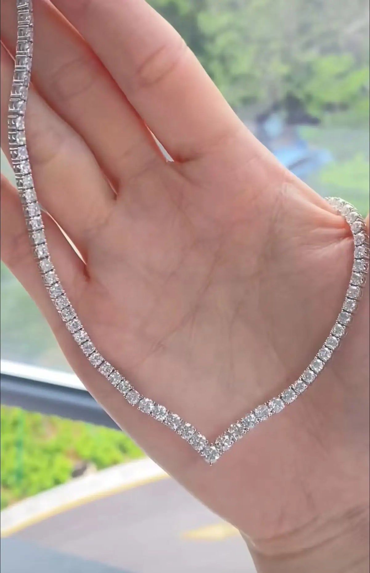 A close-up of a hand holding a silver tennis necklace paved with clear rectangular gemstones, with a blurred outdoor background visible through a window.