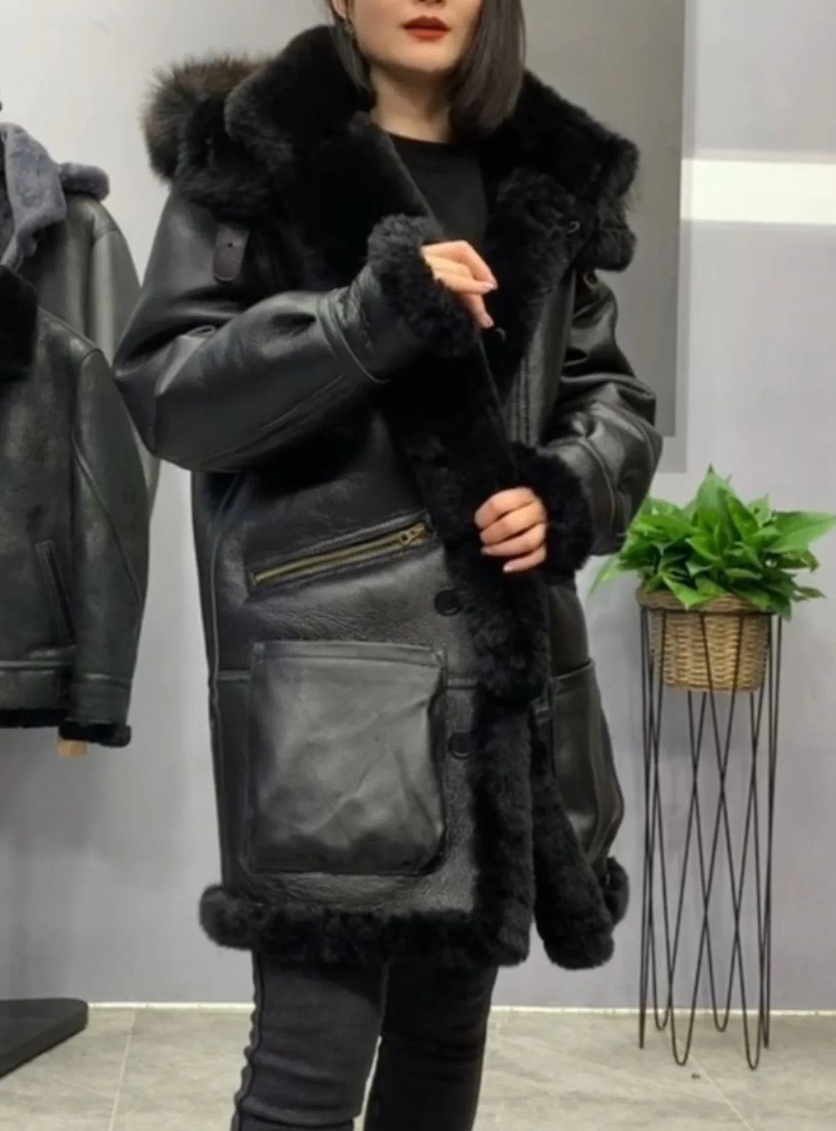 A woman wearing a black leather jacket with a large black fur collar and trim, standing in an indoor setting with a gray wall, a hanging jacket on the left, and a potted plant on the right.