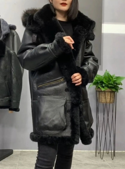 A woman wearing a black leather jacket with a large black fur collar and trim, standing in an indoor setting with a gray wall, a hanging jacket on the left, and a potted plant on the right.