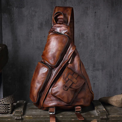 A brown distressed leather sling backpack with multiple zippered compartments and a front pocket, displayed on a rustic wooden crate against a dark gray textured background.