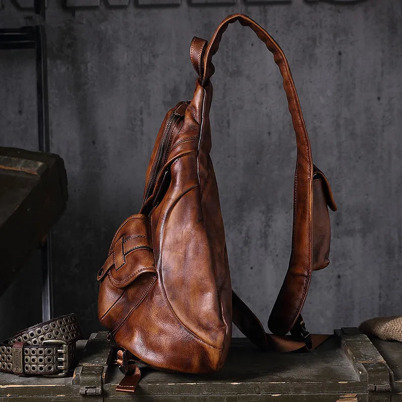 A brown leather sling backpack with a long shoulder strap, shown standing upright on a rustic wooden crate against a dark gray textured wall.