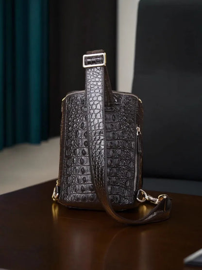 A dark brown crocodile-embossed leather crossbody bag with gold-tone hardware, resting on a dark wooden table in an indoor setting.