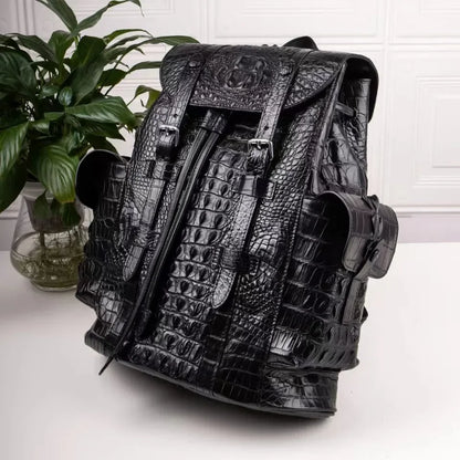 A black crocodile-embossed leather backpack with multiple front pockets and buckle details, displayed on a white surface next to a green potted plant.
