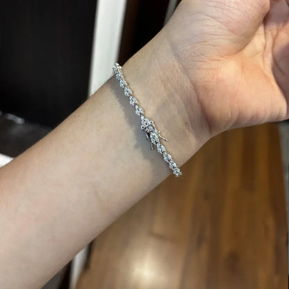 A close-up of a person's wrist wearing a silver tennis bracelet with clear gemstones, set against an indoor background with a wooden floor.