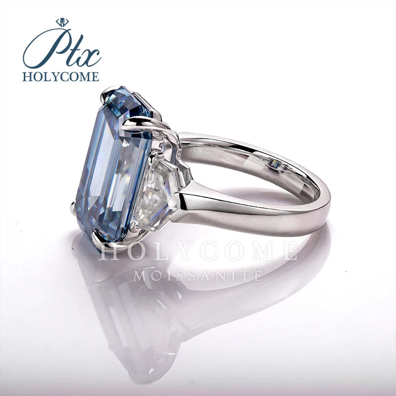 A silver-tone ring featuring a large rectangular emerald-cut light blue moissanite gemstone, shown on a reflective white surface with a brand watermark 'HOLYCOME' in the upper left.