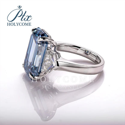 A silver-tone ring featuring a large rectangular emerald-cut light blue moissanite gemstone, shown on a reflective white surface with a brand watermark 'HOLYCOME' in the upper left.