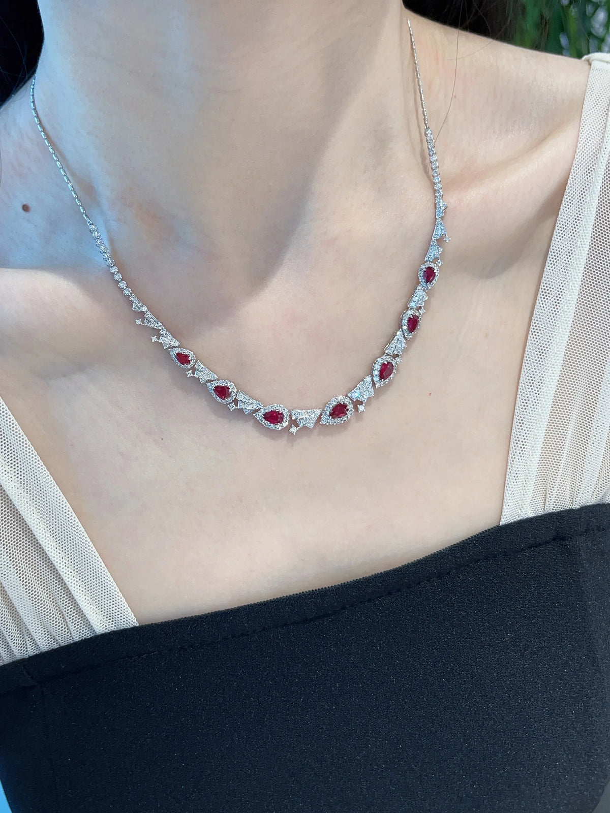 A close-up of a woman wearing a delicate silver-toned necklace with a series of red oval gemstones and small clear stones, styled with a black and white top.