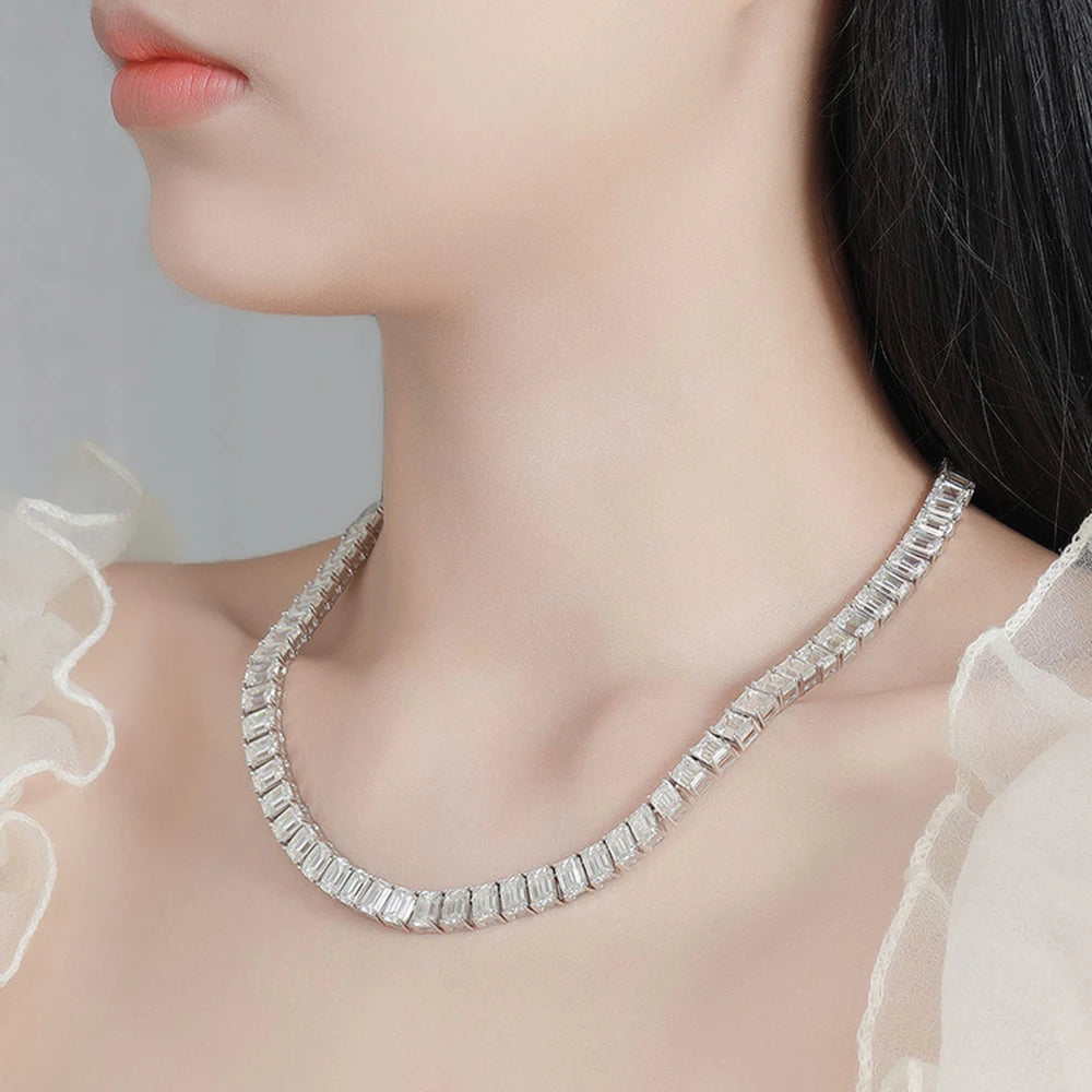 A close-up of a model wearing a silver-toned tennis-style necklace with rectangular clear stones, paired with a sheer white ruffled top.
