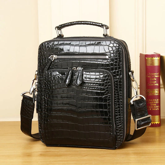 A black crocodile-embossed leather crossbody bag with a top handle, front zippered pockets, and a detachable shoulder strap, displayed on a wooden surface next to a red book.