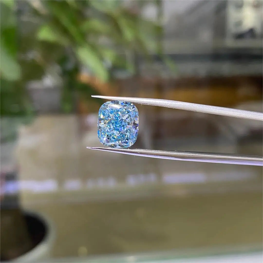 A cushion-cut blue diamond held between metal tweezers, with a softly blurred background showing green plants and warm indoor tones.
