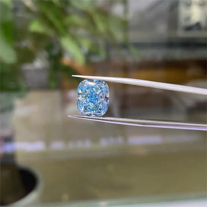 A cushion-cut blue diamond held between metal tweezers, with a softly blurred background showing green plants and warm indoor tones.