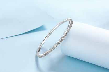 A thin silver bangle bracelet with a row of small clear stones along the front, displayed on a white cylindrical stand against a soft light blue background.