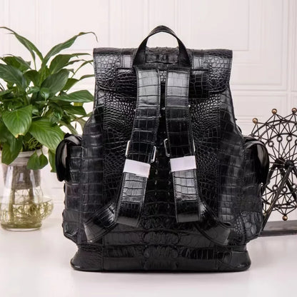 A black crocodile-embossed leather backpack with dual front straps and silver buckles, displayed on a white surface with a green plant on the left and a decorative metal sculpture on the right.