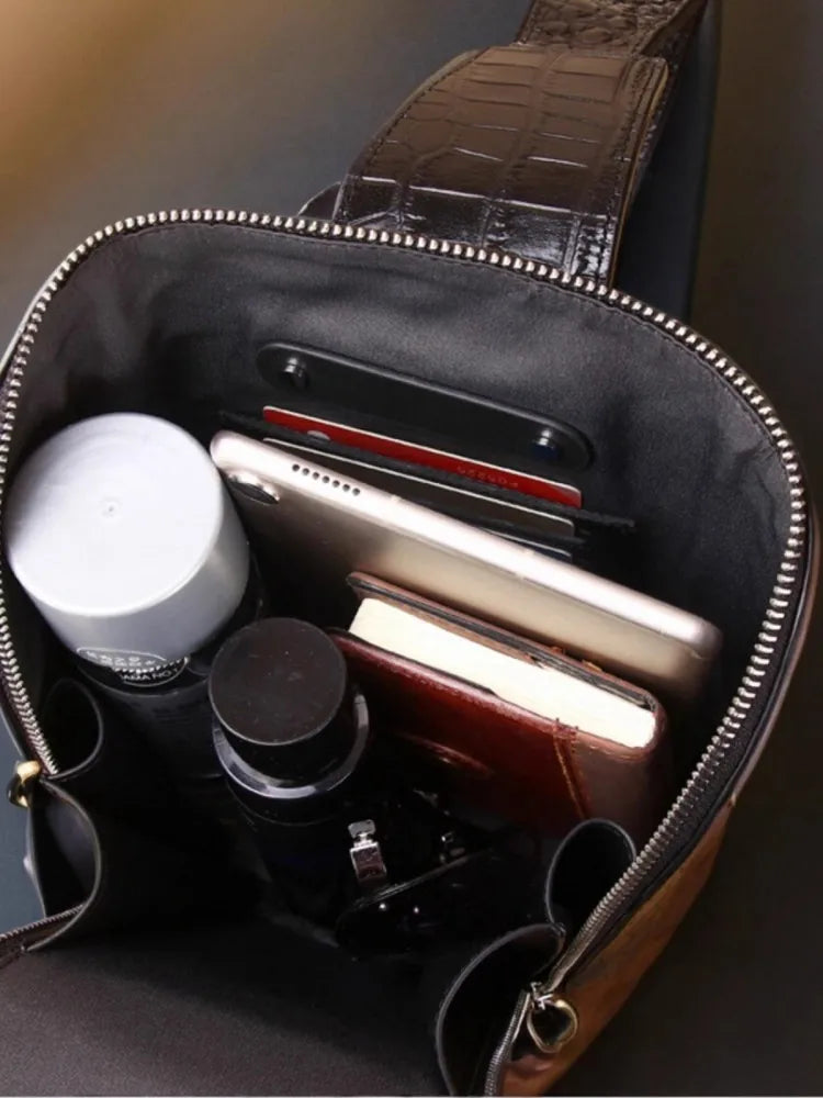 Open brown leather bag with a crocodile-embossed shoulder strap, showing a black interior organizer filled with a smartphone, wallet, notebook, and small accessories.