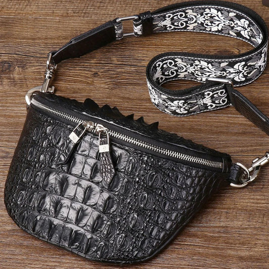 Black crocodile-embossed faux leather fanny pack with a wide black and white patterned strap, resting on a wooden surface.