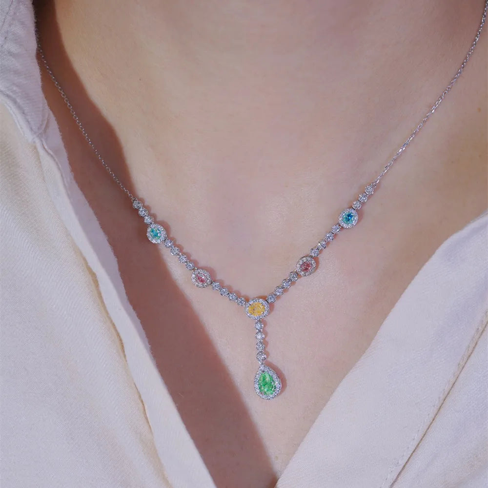 A close-up of a model wearing a silver Y-shaped necklace adorned with multiple colorful gemstones, including a prominent green teardrop pendant, a yellow center stone, and smaller blue, pink, and green stones.