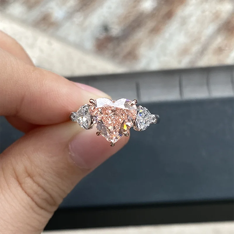 A close-up of a hand holding a rose gold ring with a heart-shaped center stone and clear accent stones, set against a dark gray and beige blurred background.