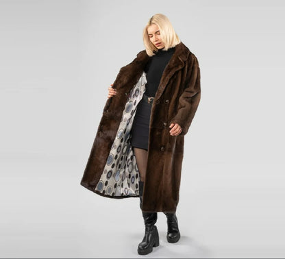 A blonde model wearing a long brown faux fur coat with a patterned lining, standing against a light gray studio background.