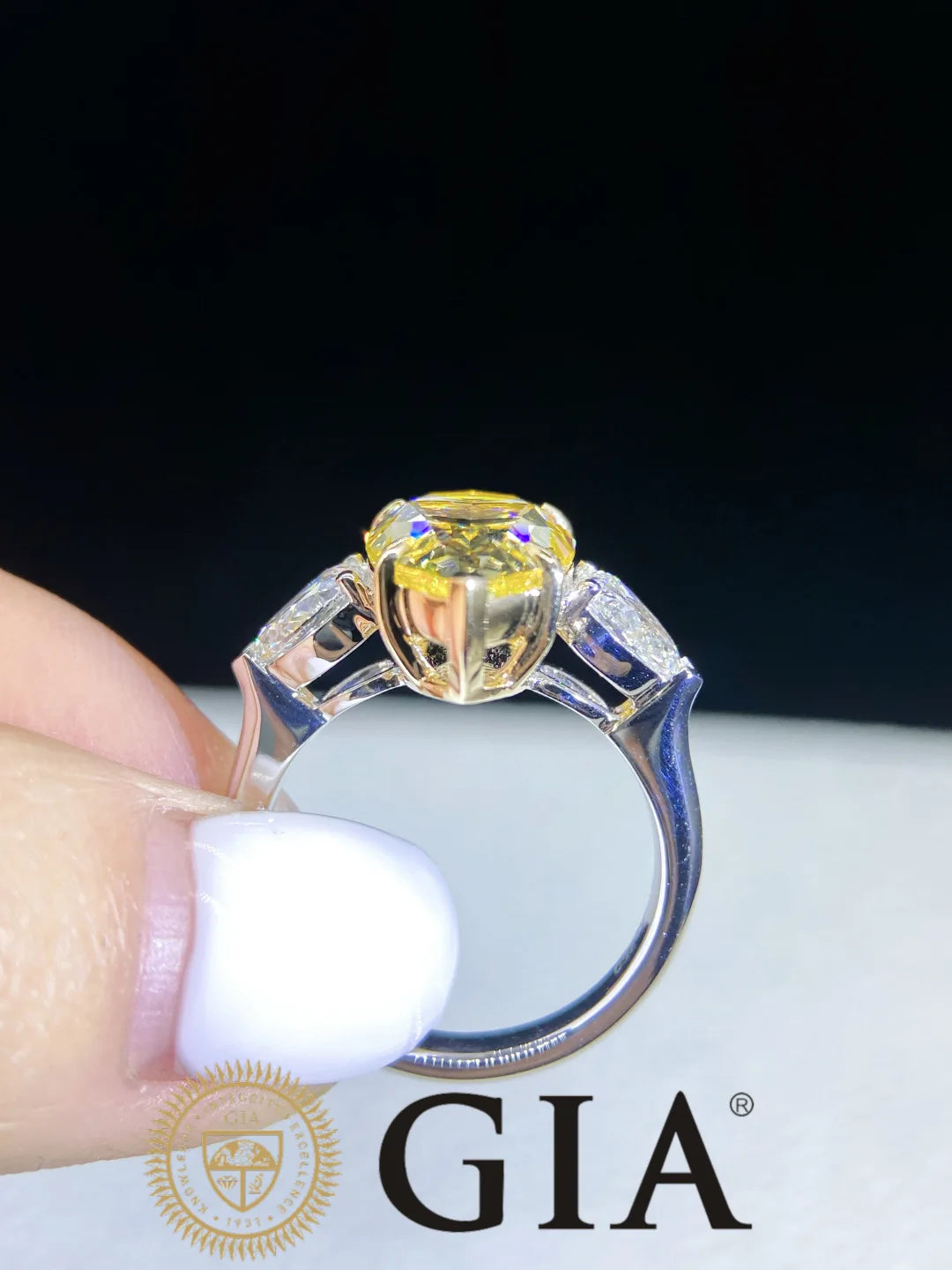 A close-up of a yellow gemstone ring held between fingers against a dark background, with a GIA logo watermark at the bottom.