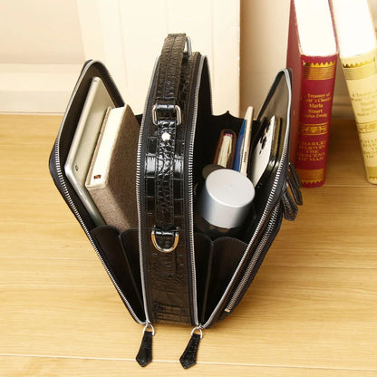 A black crocodile-embossed zip-around organizer bag shown open on a light wood tabletop, holding notebooks, a small bottle, and other accessories, with books in the background.