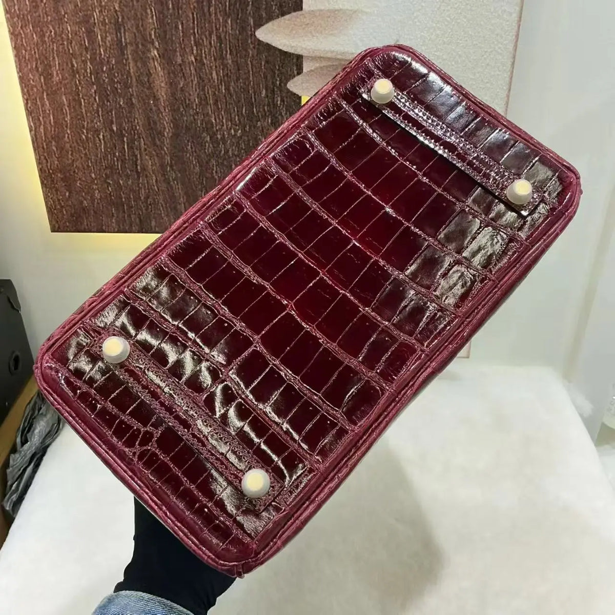 A glossy crocodile-embossed leather handbag shown from the bottom, held by a hand. The bag is a deep burgundy red with four cream-colored round studs on the bottom corners.