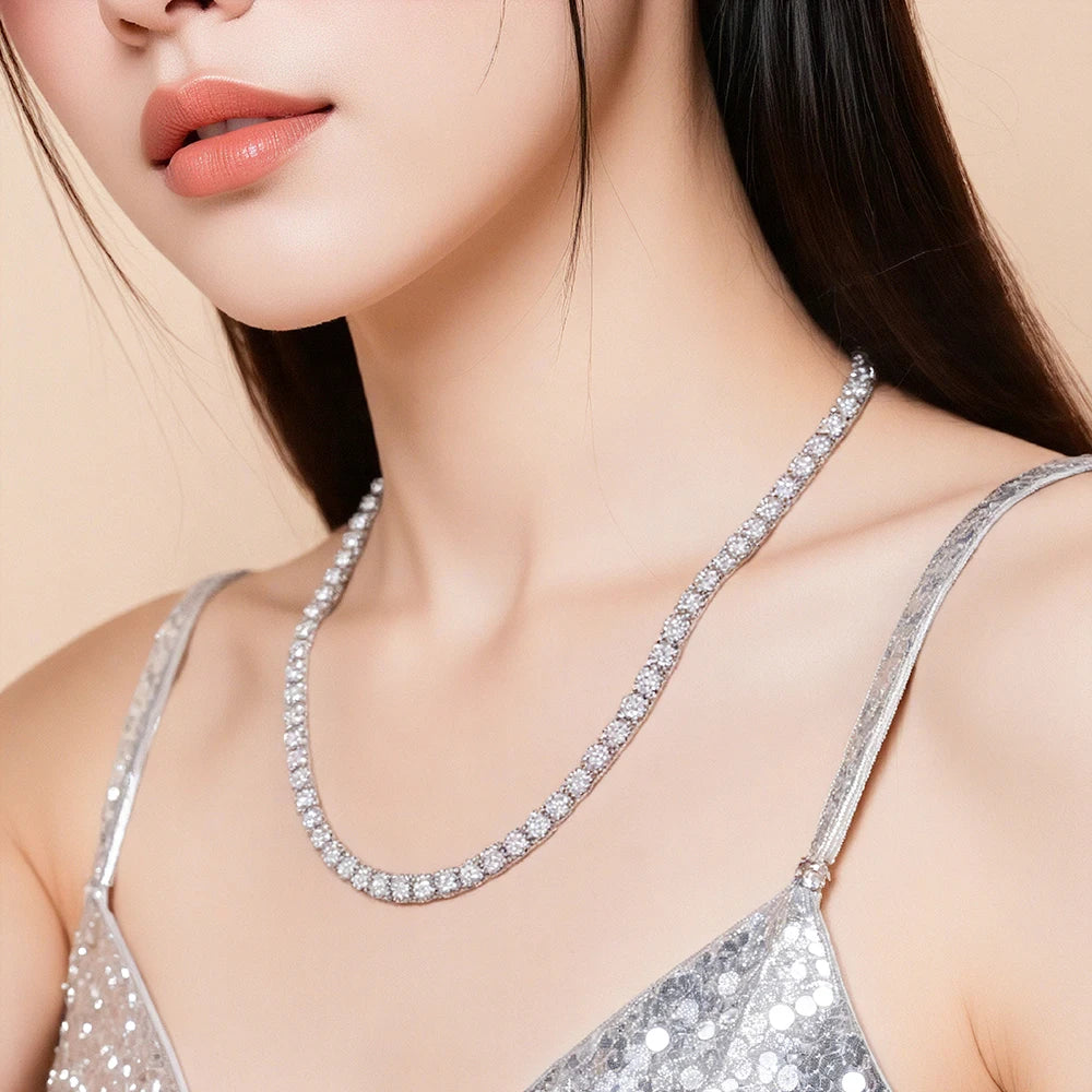 A close-up of a model wearing a sparkling silver tennis-style necklace with clear stones, paired with a silver sequined top against a soft beige background.