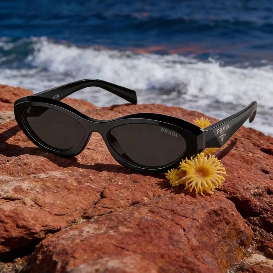 Prada cat-eye sunglasses with black frames and dark lenses resting on a red rock with ocean waves in the background.