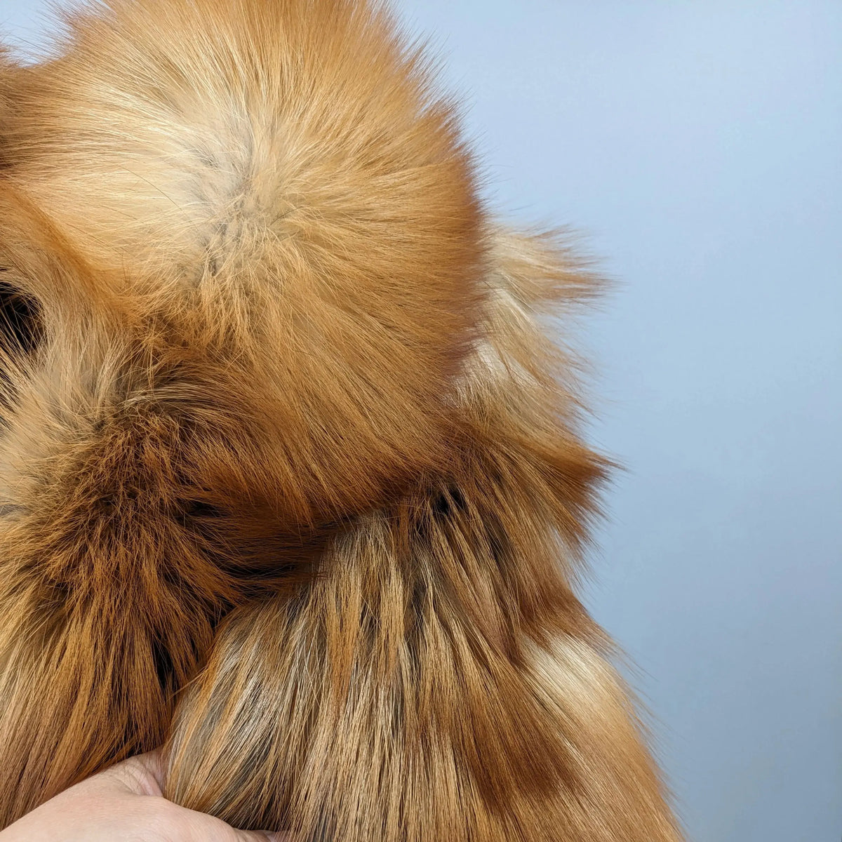 Close-up of a fluffy faux fur or sheepskin material in warm tan and brown tones, with a hand holding it against a pale blue background.