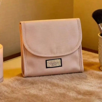 A light pink Gucci cosmetic pouch with a flap closure and a black and white logo patch, resting on a beige furry surface.