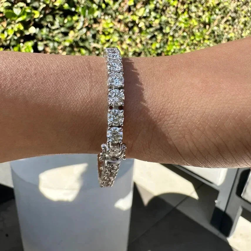 A silver-toned diamond tennis bracelet worn on a person's wrist, photographed outdoors with green foliage and a white cylindrical pedestal in the background.
