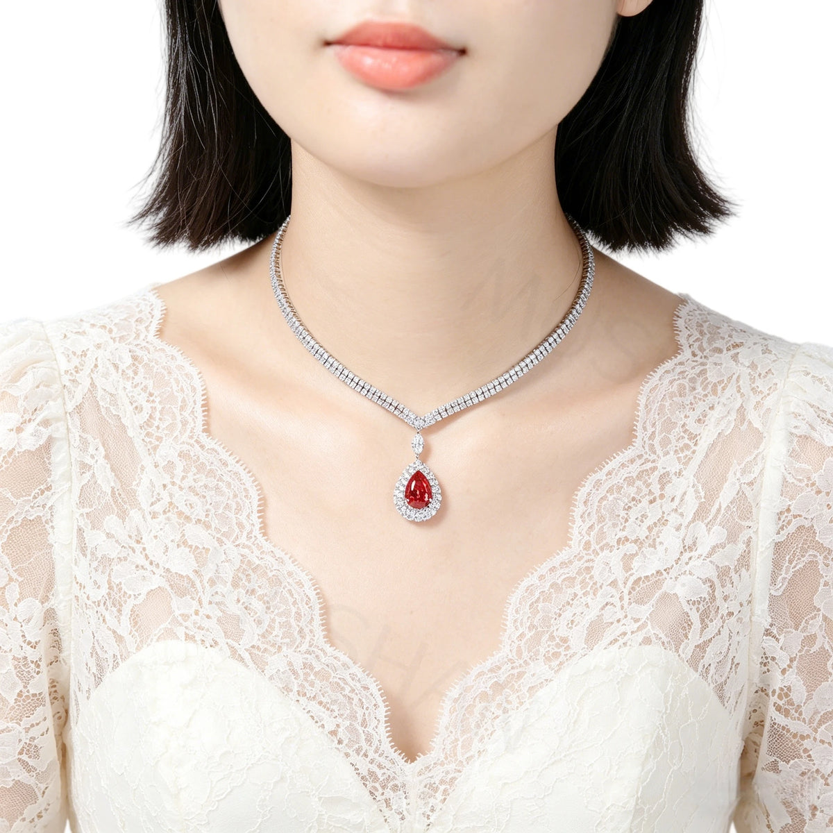 A close-up of a model wearing a silver-toned diamond-studded necklace with a teardrop-shaped red gemstone pendant, paired with a white lace top against a clean white background.