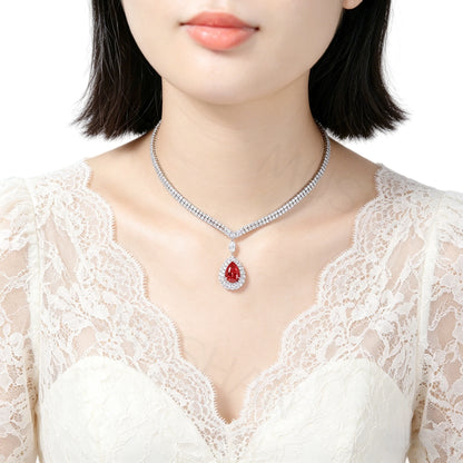 A close-up of a model wearing a silver-toned diamond-studded necklace with a teardrop-shaped red gemstone pendant, paired with a white lace top against a clean white background.