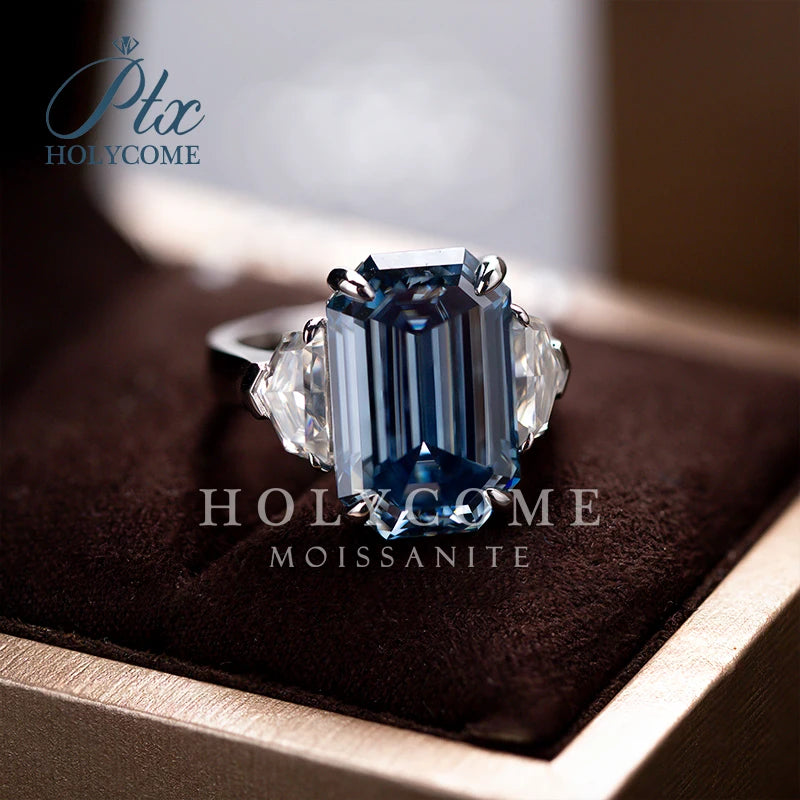 A halo Moissanite ring featuring a large rectangular blue moissanite center stone with clear baguette side stones, set in a silver-toned band and displayed on a dark brown velvet ring box. The brand logo 'Ptc HOLYCOME' is visible in the top left corner.