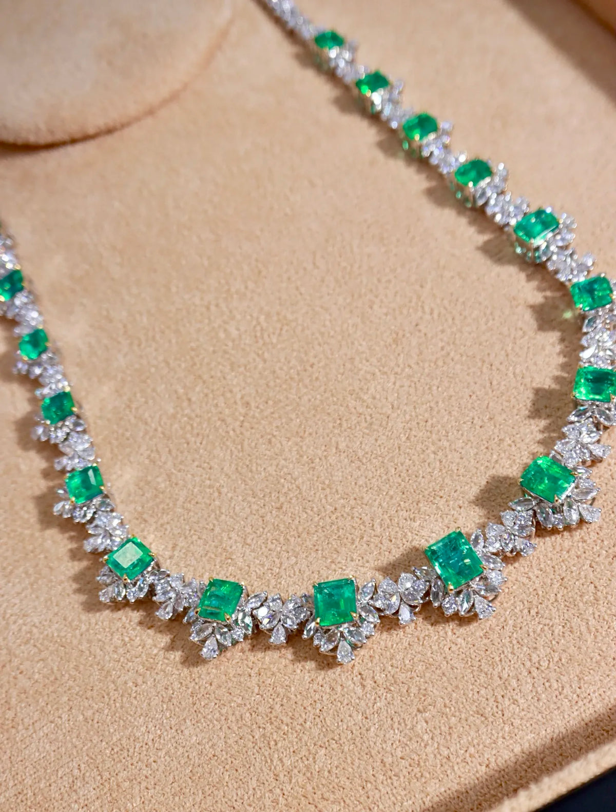 A close-up of a necklace featuring bright green emerald-cut gemstones and silver-toned floral settings, displayed on a beige textured surface.