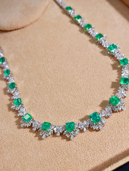 A close-up of a necklace featuring bright green emerald-cut gemstones and silver-toned floral settings, displayed on a beige textured surface.