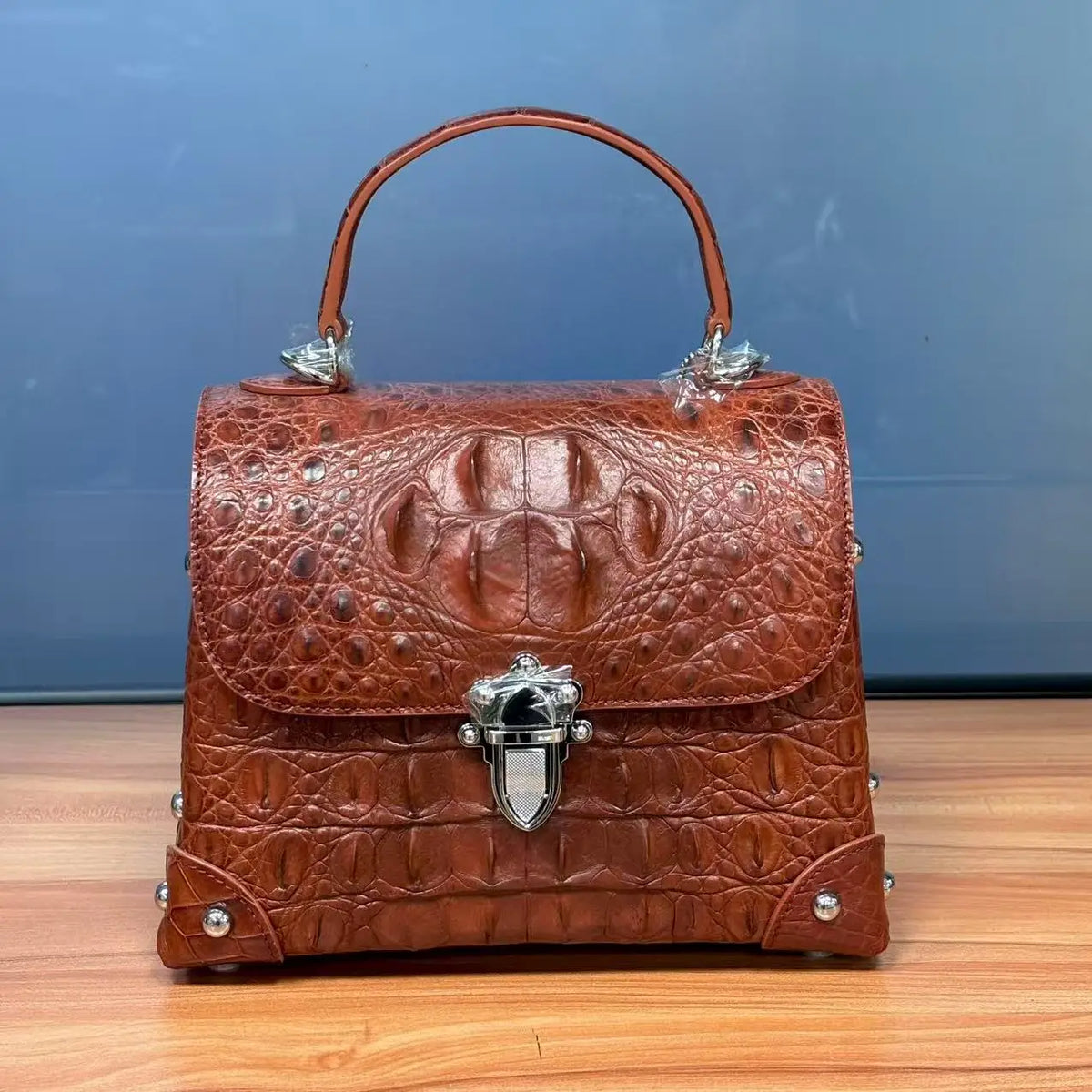 A brown crocodile-embossed leather handbag with a top handle, silver-tone hardware, and a decorative clasp, displayed on a wooden surface against a blue background.